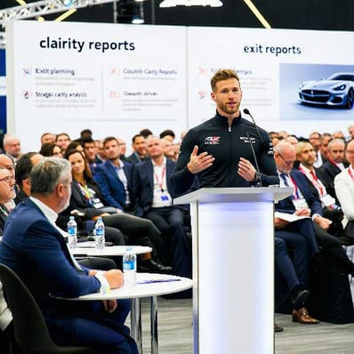 The image depicts a bustling Formula 1 conference setting with attendees engaged in discussions and networking In the foreground an attractive 40 year old driver stands confidently at a podium gesturing passionately as they share insights about exit-1